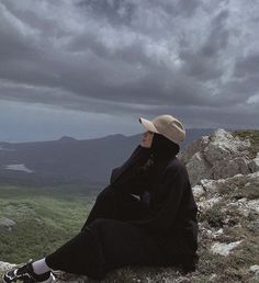 a person sitting on top of a mountain with a hat and black jacket over their head