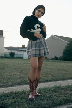 a woman standing in the grass with her legs crossed wearing boots and a sweater over her shoulders