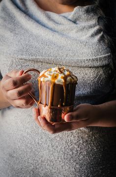 a woman holding a cup of hot chocolate with whipped cream on top and caramel drizzle