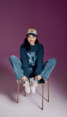 a woman sitting on top of a wooden chair wearing jeans and a baseball cap smiling at the camera