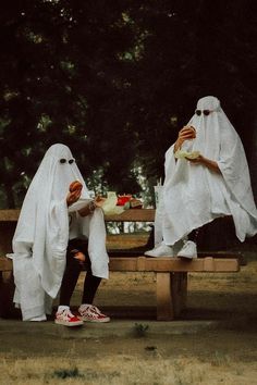 two people dressed in ghost costumes sitting on a park bench eating sandwiches and drinking juice