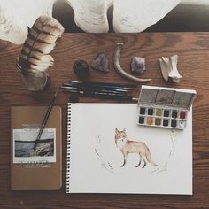 a desk topped with art supplies next to a painting on top of a wooden table