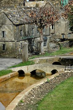 an old stone bridge over a small pond in front of some buildings with windows on each side