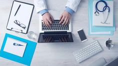 two hands typing on a laptop keyboard next to a phone and other office supplies, including a doctor's stethoscope
