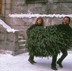 a man and woman walking in the snow carrying a christmas tree on their back,
