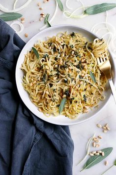 a white bowl filled with pasta and topped with olives, pine nuts and sage