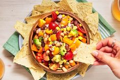 a person holding a tortilla chip in front of a bowl filled with fruit and veggies