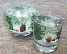 two glass candles sitting on top of a wooden table covered in snow and pine cones