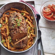 a pan filled with meat and french fries on top of a wooden table next to a bowl of tomatoes