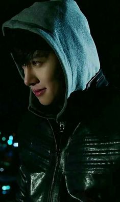 a young man wearing a leather jacket and hoodie at night with city lights in the background