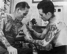 two men with tattoos on their arms looking at an old fashioned camera and one is holding something in his hand
