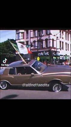 an old car driving down the street with a flag on it's roof and some buildings in the background