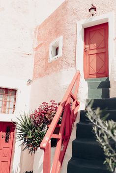 a red door is on the side of a white building with plants and flowers in front