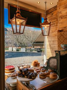 a table filled with pastries and donuts under two hanging lights in front of a window