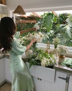 a woman in a green dress arranging flowers on a window sill next to a sink