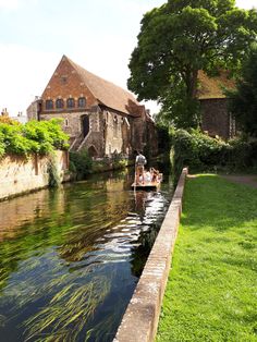 two people are on a boat in the water next to a brick building and green grass
