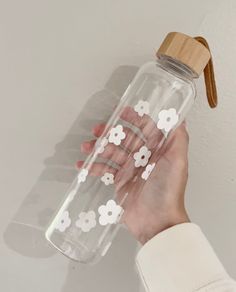 a hand holding a clear water bottle with white flowers on the side and a wooden lid