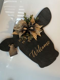 a welcome sign hanging on the side of a white wall with leaves and flowers in it