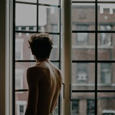 a shirtless man standing in front of a window looking out at the city outside