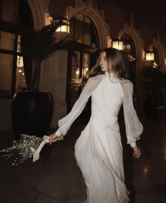 a woman in a long white dress holding a bouquet of flowers and walking away from the camera