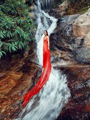 a woman in a long red dress standing by a waterfall