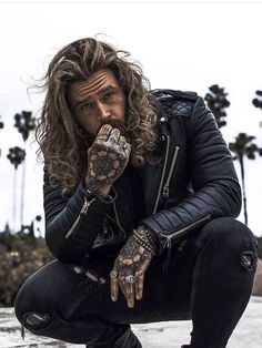 a man with long hair and tattoos on his hands crouching down in front of palm trees