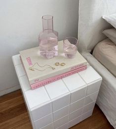 two pink glasses sitting on top of a white box next to a bed and nightstand