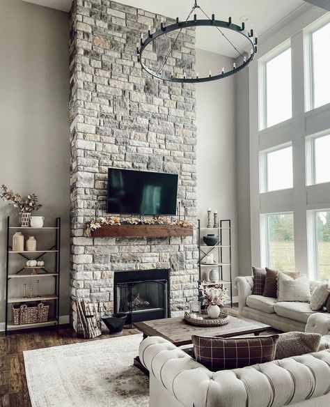 A flat panel TV is mounted to a gray stone two story fireplace above a stained wood mantel. The TV faces a light gray Chesterfield sofa and wood coffee table lit by a black wagon wheel chandelier. Instagram, Country, Living Room Grey, Two Story Fireplace, White Shiplap, Tall Fireplace, Grey Stone Fireplace, White Shiplap Wall, Grey Fireplace