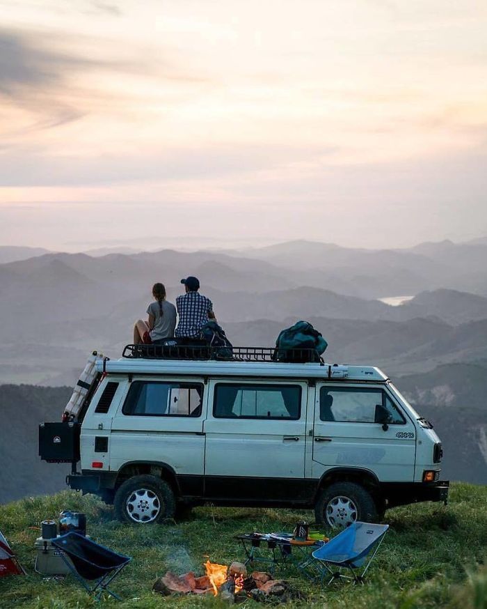 two people sitting on top of a van in the mountains with their campervan
