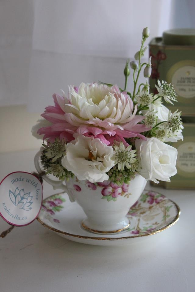 a white and pink flower arrangement in a teacup on a saucer next to two boxes