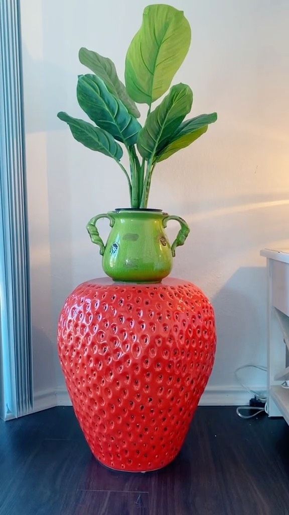 a green potted plant sitting on top of a red strawberry shaped vase next to a white wall