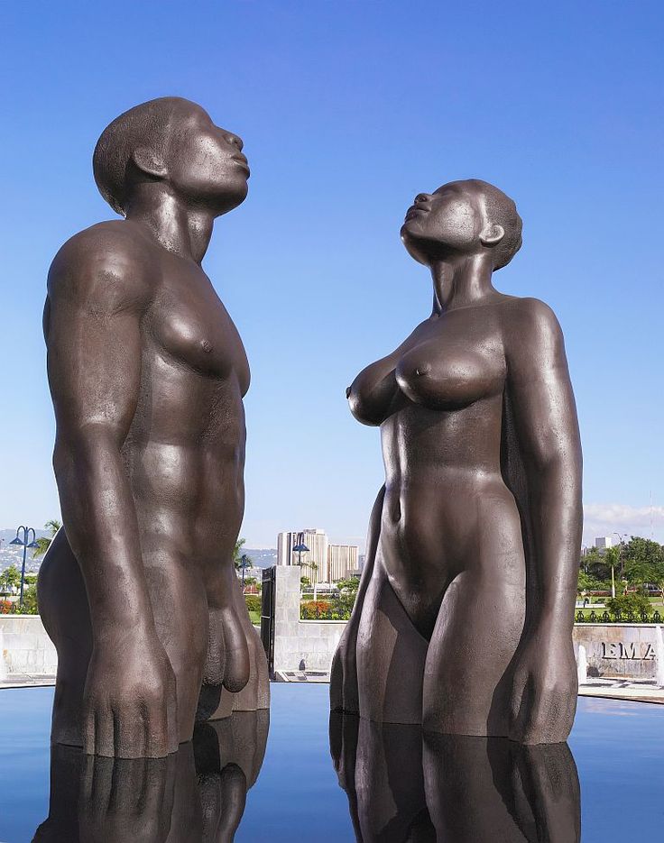 two statues are standing next to each other in front of a body of water with buildings in the background