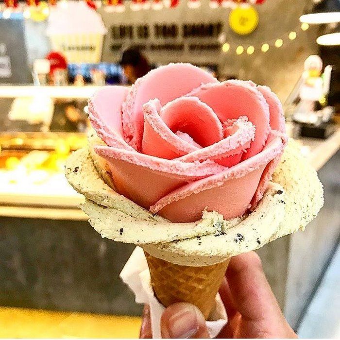 a person holding an ice cream cone with pink frosting on it in front of a food court