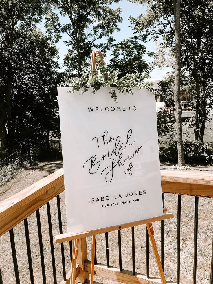 a welcome sign on an easel in front of a wooden fence with trees behind it