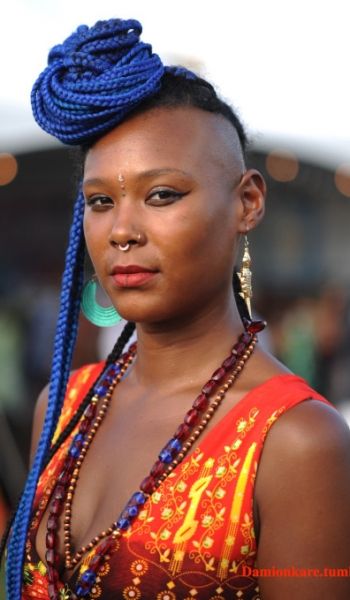 a woman with blue braids in her hair and wearing an orange top is looking at the camera
