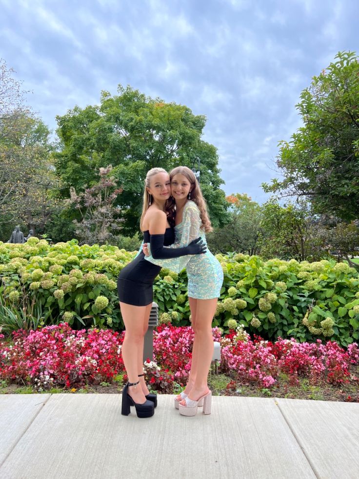 two young women standing next to each other in front of flowers and bushes with their arms around each other