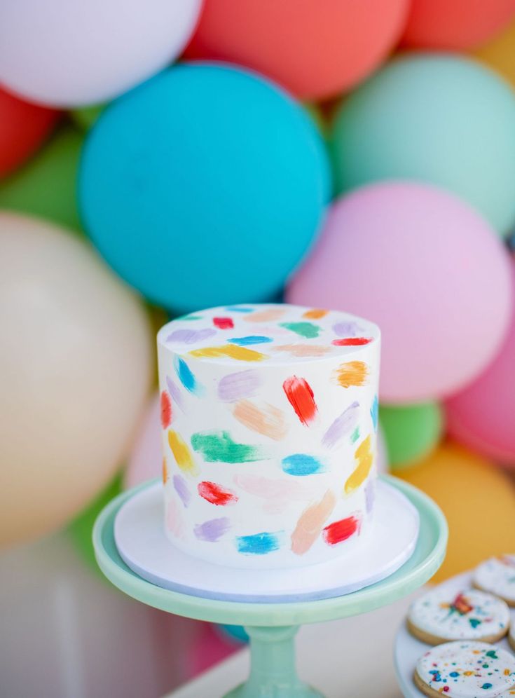 there is a colorful cake and cookies on the table with balloons in the back ground