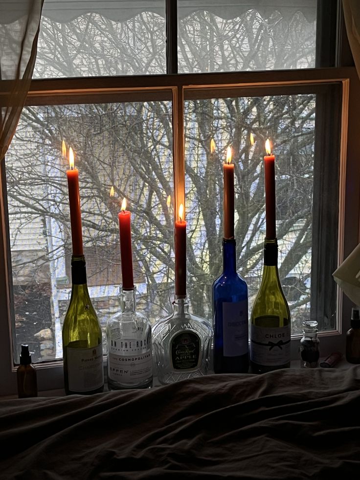 three wine bottles are sitting in front of a window, with candles lit on the windowsill