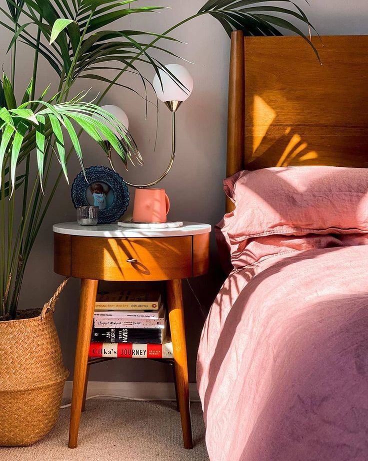 a bed with pink sheets and pillows next to a wooden table with a plant on it