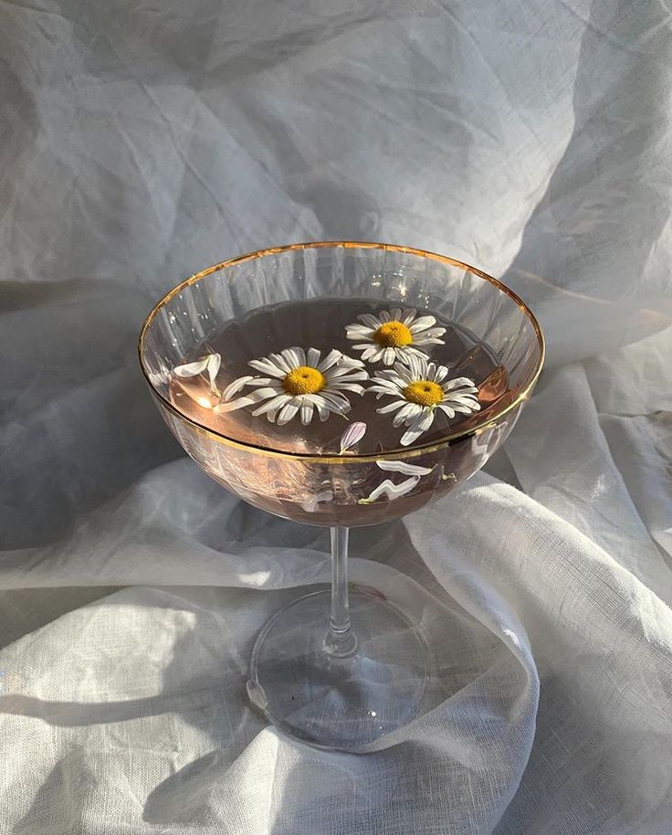 a glass filled with liquid and daisies on top of a white cloth covered table