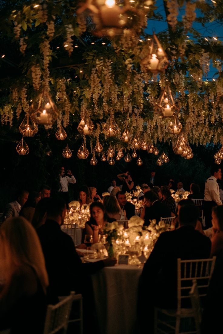 a group of people sitting around a dinner table with lights hanging from the ceiling above them