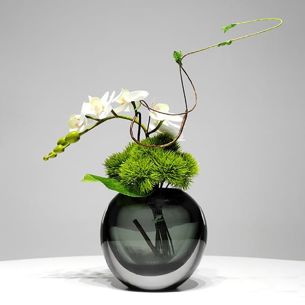 a black vase with white flowers and green grass in it sitting on a table next to a gray wall