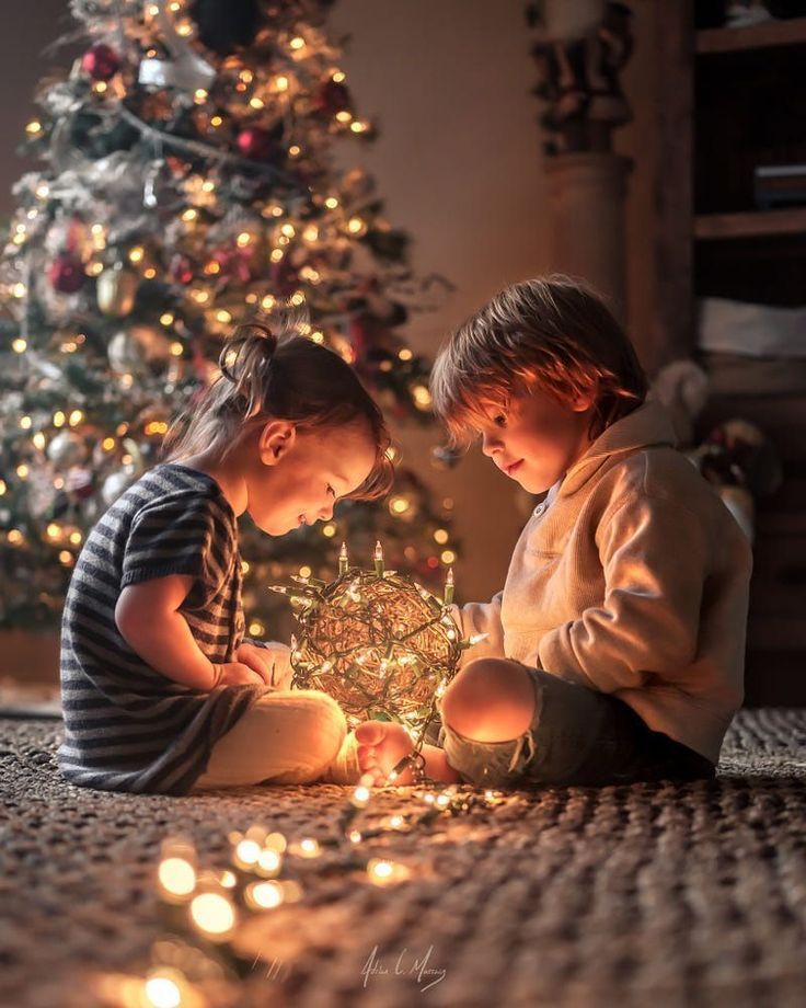two children sitting on the floor looking at a christmas ornament in front of a christmas tree