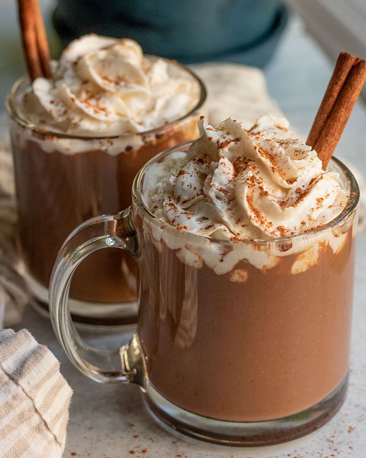 two mugs filled with hot chocolate and topped with whipped cream, cinnamon sticks and powdered sugar