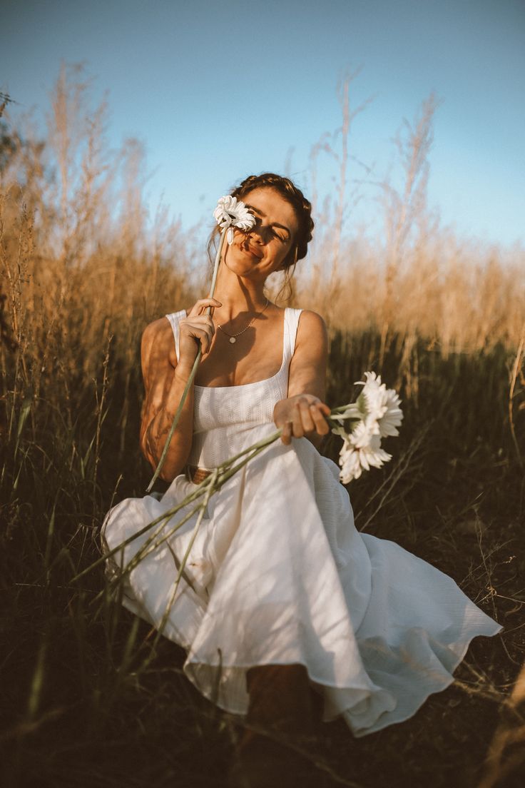 a woman in a white dress holding a flower and looking at the camera while standing in tall grass
