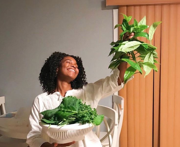 a woman holding up a bowl filled with green leafy vegetables on top of a table