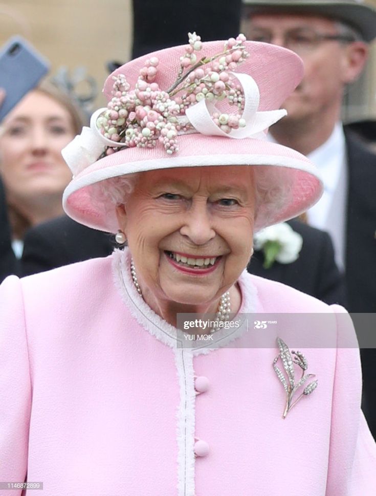 an older woman wearing a pink outfit and hat with flowers on it, standing in front of other people