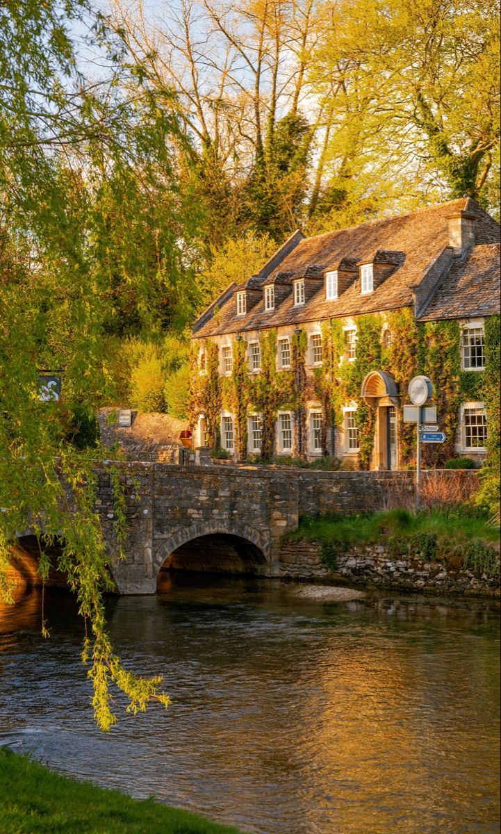 A period property in the evening sunshine behind a river and small road bridge. The spring scene is surrounded by trees that are beginning to bloom. Urban, Country, Destinations, London England, Hotels, Arlington Row, Countryside Hotel, The Cotswolds England, Cotswolds England