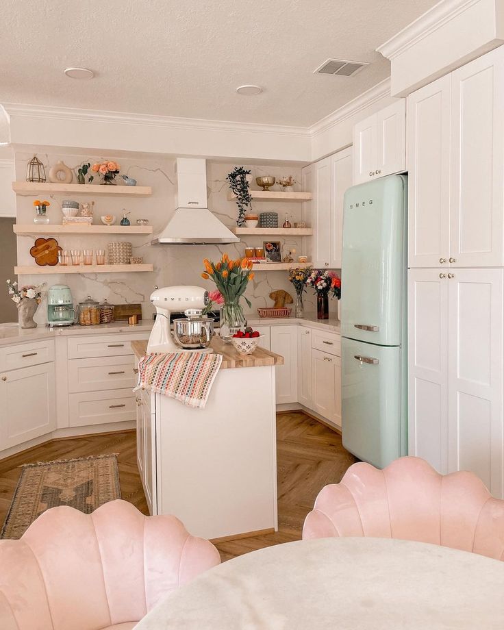 a white kitchen with pink chairs and an old fashioned refrigerator in the center, surrounded by open shelving