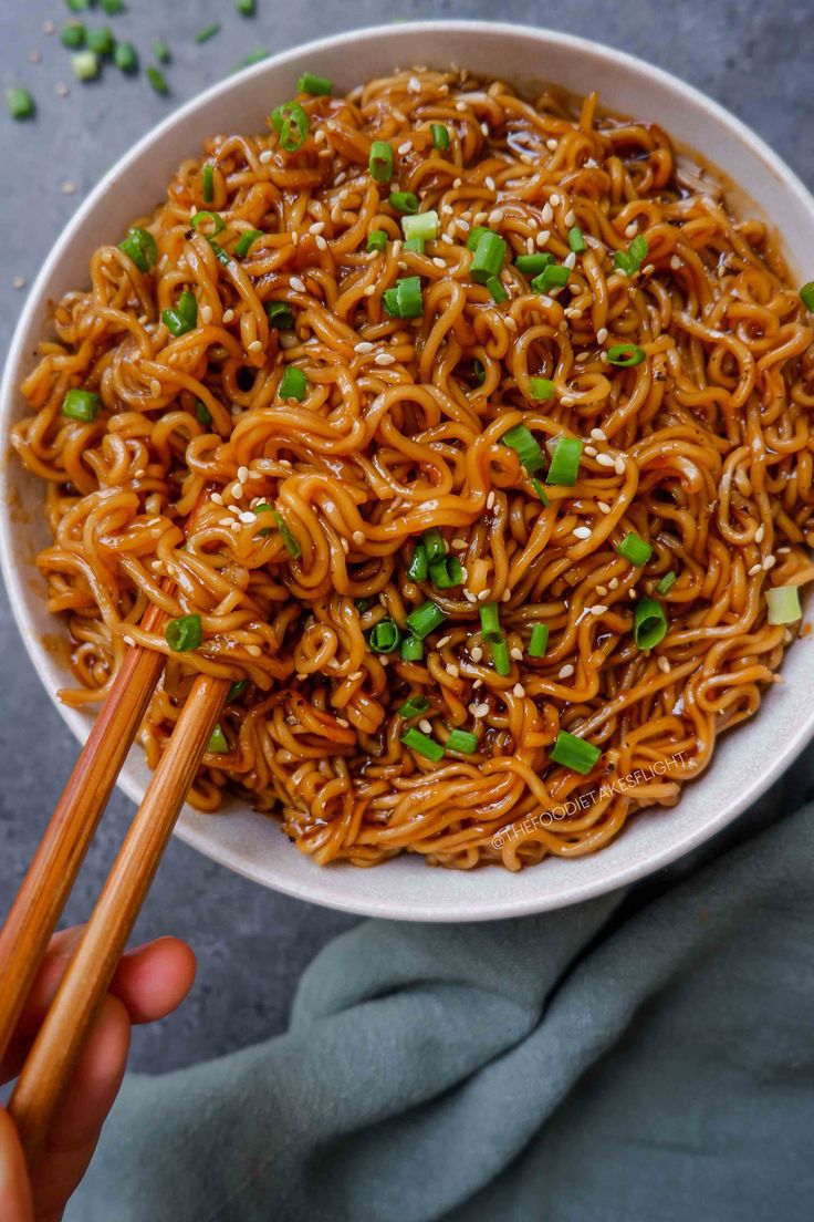 someone holding chopsticks over a bowl of noodles with sesame seeds and scallions